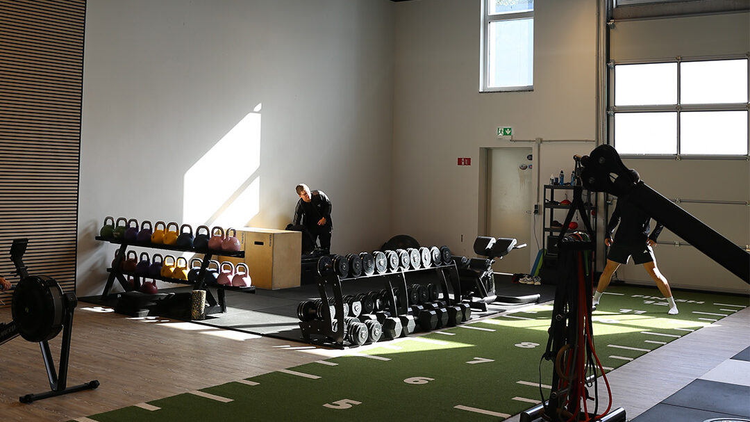 Trainingshalle 2 mit Sprintbahn, Hantelablagen und Sektionaltor, im Hintergrund trainierende Personen.